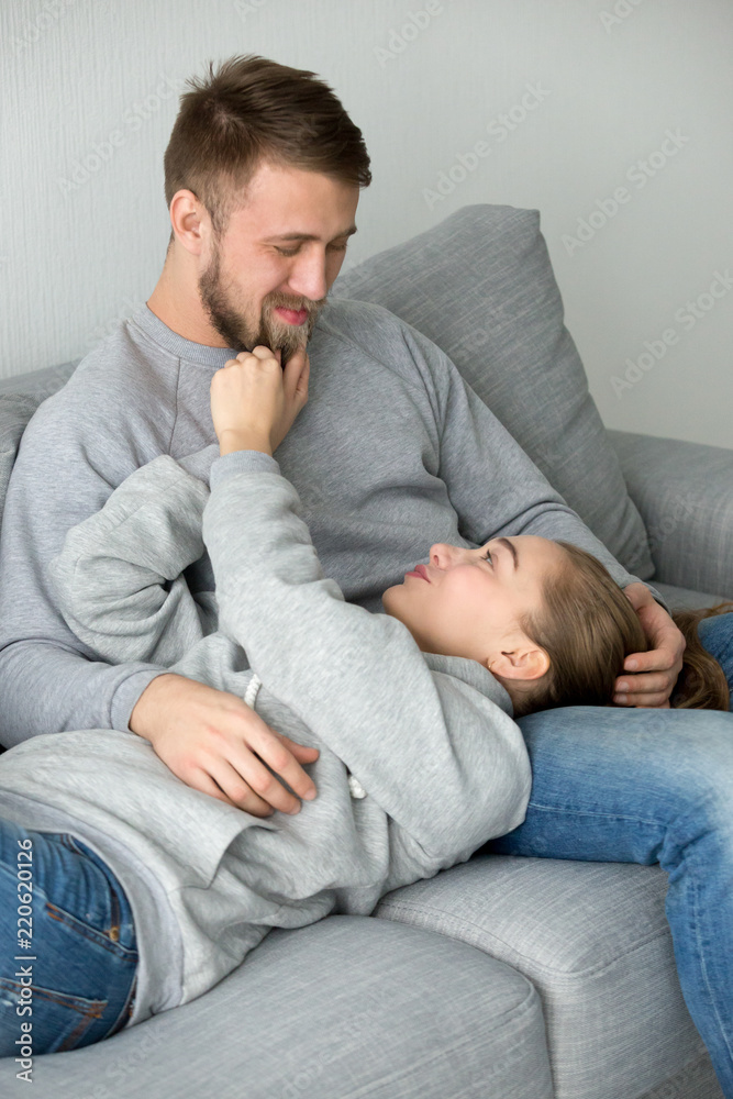 Loving young girlfriend lying on boyfriend knees caress him playing with beard, happy woman relax in man arms looking in eyes, millennial couple having fun resting on cozy couch at home hugging
