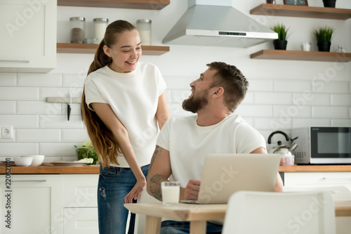 Smiling husband and wife looking at each other before having breakfast at home, man using laptop in kitchen, talking to happy girlfriend, young excited couple enjoy romantic morning together