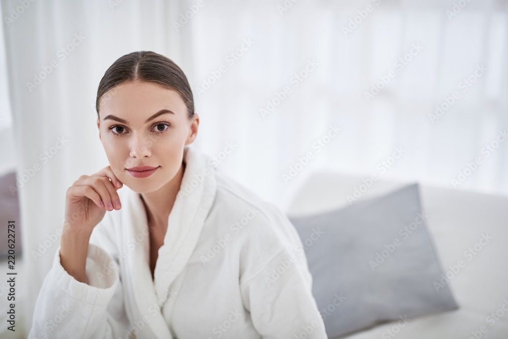 Perfect skin. Portrait of lovely girl in white soft bathrobe at beauty salon. She touching chin and looking at camera with smile Perfect skin. Portrait of lovely girl in white soft bathrobe at beauty salon. She touching chin and looking at camera with smile