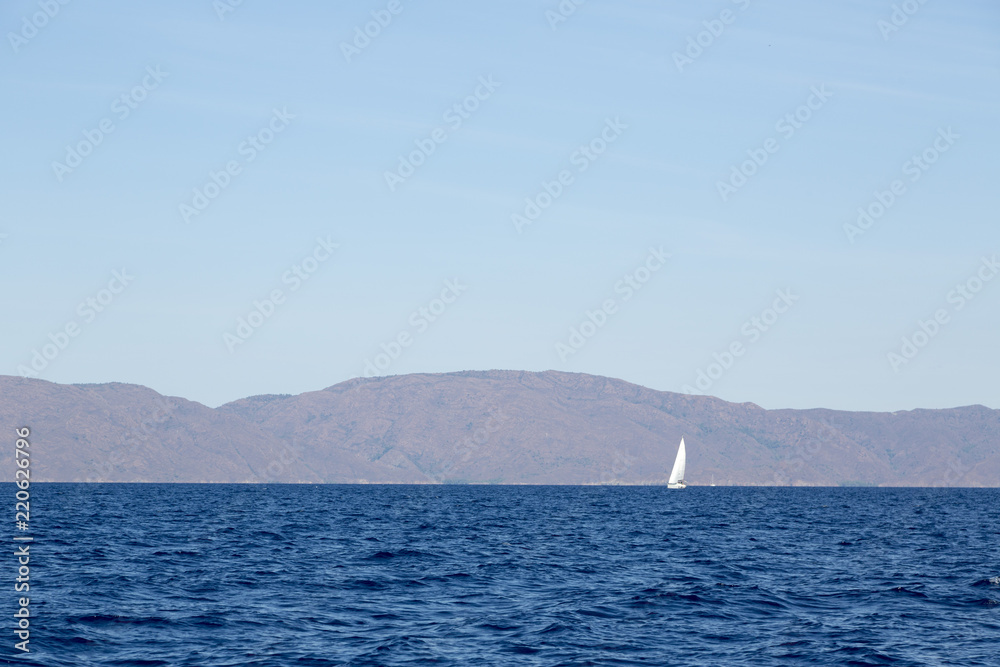 Fototapeta premium Sailing boat cruising in blue waters, mountains and shore seeing in background