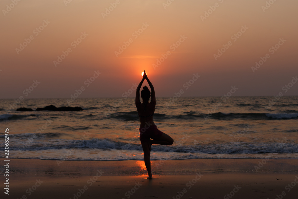 Yoga teacher doing yoga at sunset on the beach. Pose on balance, pose tree. Hands are raised and connected together. Natural backlight.