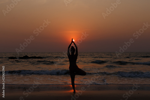Young slender girl with her back doing yoga at sunset on the beach. Pose on balance, pose tree. Hands are raised and connected together. Natural backlight.