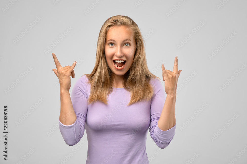 Cheerful young blonde girl smiles and shows a hand sign rock ' n ' roll. The concept of fun at a party, heavy music, rock. Studio isolated portrait on grey background Cheerful young blonde girl smiles and shows a hand sign rock ' n ' roll. The concept of fun at a party, heavy music, rock. Studio isolated portrait on grey background