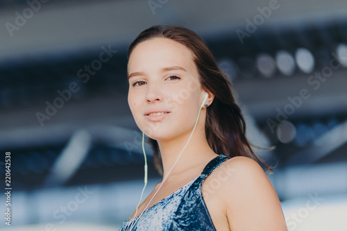 Close up shot of good looking healthy European woman listens music with unrecognizable device, looks directly at camera, poses against blurred background. People, youth, entertainmet concept photo