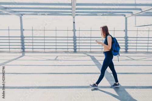 Horizontal shot of good looking sportswoman dressed in casual outfit, carries rucksack, being addicted to modern technologies, always in touch, listens favourite song. Active lifestyle concept