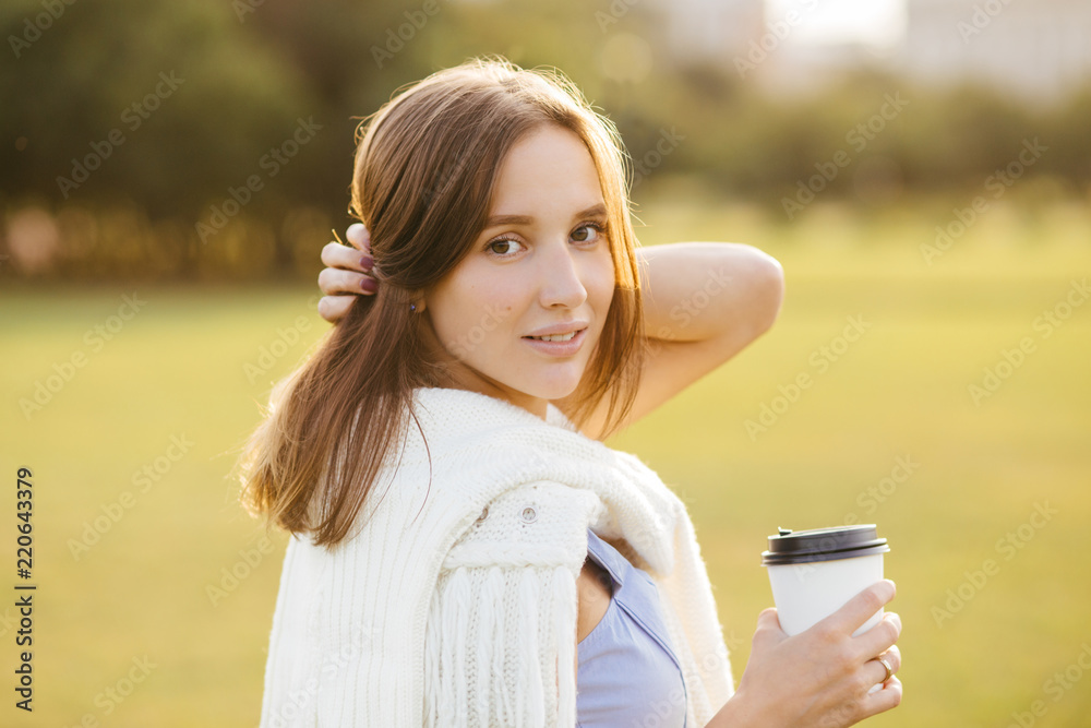 Portrait of beautiful pretty female with dark hair, healthy pure skin, dressed in casual outfit, drinks takeaway coffee, poses outside, enjoys calm atmosphere, has weekend or day off. Rest concept