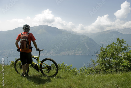 man with electric bicycle, e-bike, ebike, high mountains, observes horizon, valley and lake of Como, forest, summer, sport, adventure, cloud, alps, Italy
