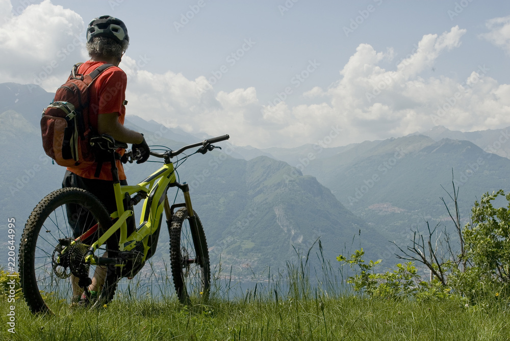 Naklejka premium man with electric bicycle, e-bike, ebike, high mountains, observes horizon, valley, lake of Como, forest, summer, sport, adventure, cloud, sun, alps, Lombardy, technology, Italy