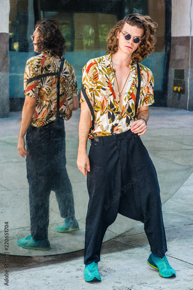 Young Man with brown curly hair, wearing round sunglasses, colorful  patterned short sleeve shirt, baggy loose pants with suspenders, patterned  sneakers, hanging old key as necklace, standing by mirror Stock Photo
