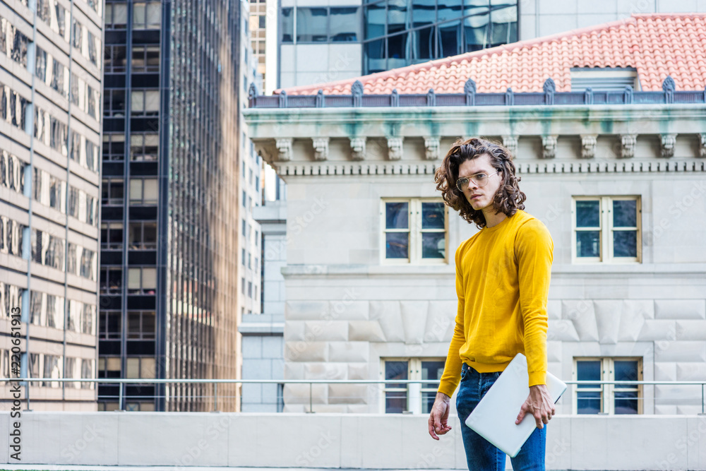 Young Hispanic American College Student Studying in New York City, with brown curly hair, wearing glasses, yellow long sleeve T shirt, blue jeans, holding laptop computer, standing outside on campus