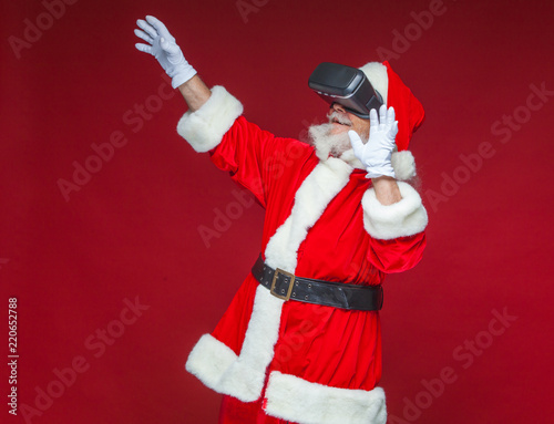 Christmas. Santa Claus in black virtual reality glasses makes gestures with his hands. Surprise, emotion. New technology. Isolated on red background.