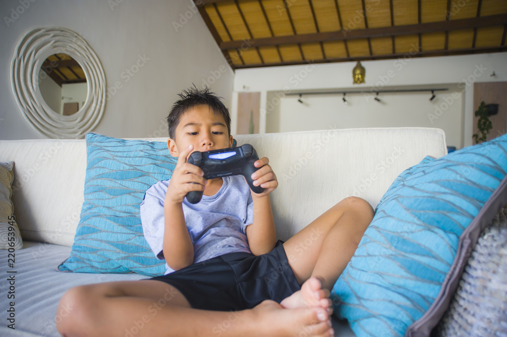  Latin young kid 8 years old excited and happy playing video game online holding remote controller enjoying having fun on couch in child gaming addiction