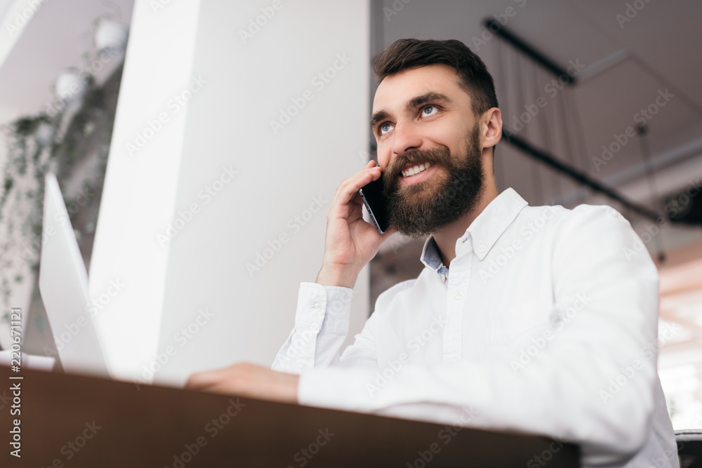 Fototapeta premium Successful handsome businessman using laptop computer and smartphone for his startup project, sitting in modern office. Stylish bearded man talking on mobile about business deals with partner