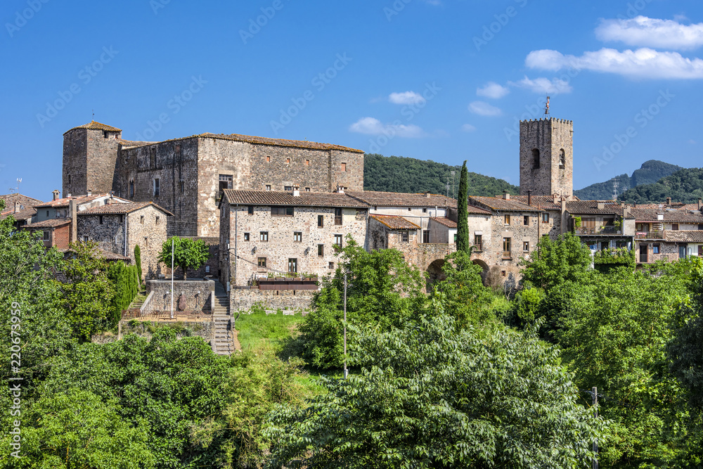 Spain, Catalonia, Santa Pau: Panoramic view on the famous skyline of old ancient fortified Spanish town with tower, houses, green trees and blue sky in the background - concept travel history.