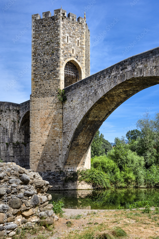 Spain, Catalonia, Besalu: Panorama view on the famous skyline of old ancient fortified Spanish town with bridge, river EL Fluvia, tower, green trees and blue sky in the background - concept history.