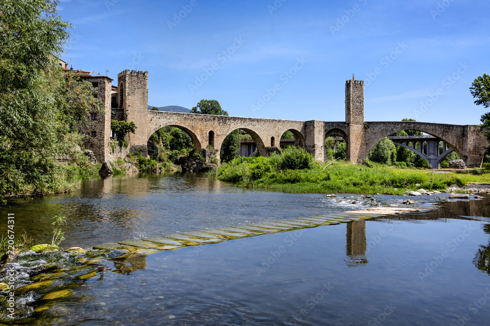 Spain, Catalonia, Besalu: Panorama view on the famous skyline of old ancient fortified Spanish town with medieval bridge, river El Fluvia, tower, houses, green trees and blue sky in the background.