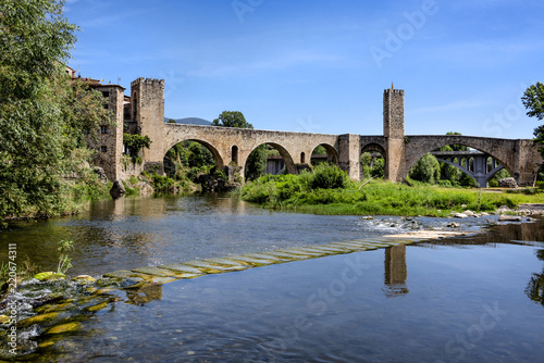 Spain, Catalonia, Besalu: Panorama view on the famous skyline of old ancient fortified Spanish town with medieval bridge, river El Fluvia, tower, houses, green trees and blue sky in the background.