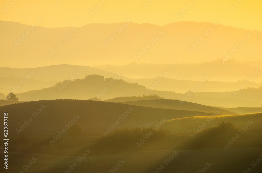 A beautiful Italian traditional rural landscape with autumn fields in the hills and a farmhouse with cypress and olive trees in a foggy morning at dawn