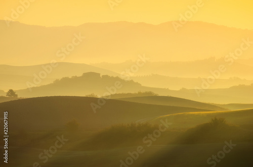 A beautiful Italian traditional rural landscape with autumn fields in the hills and a farmhouse with cypress and olive trees in a foggy morning at dawn