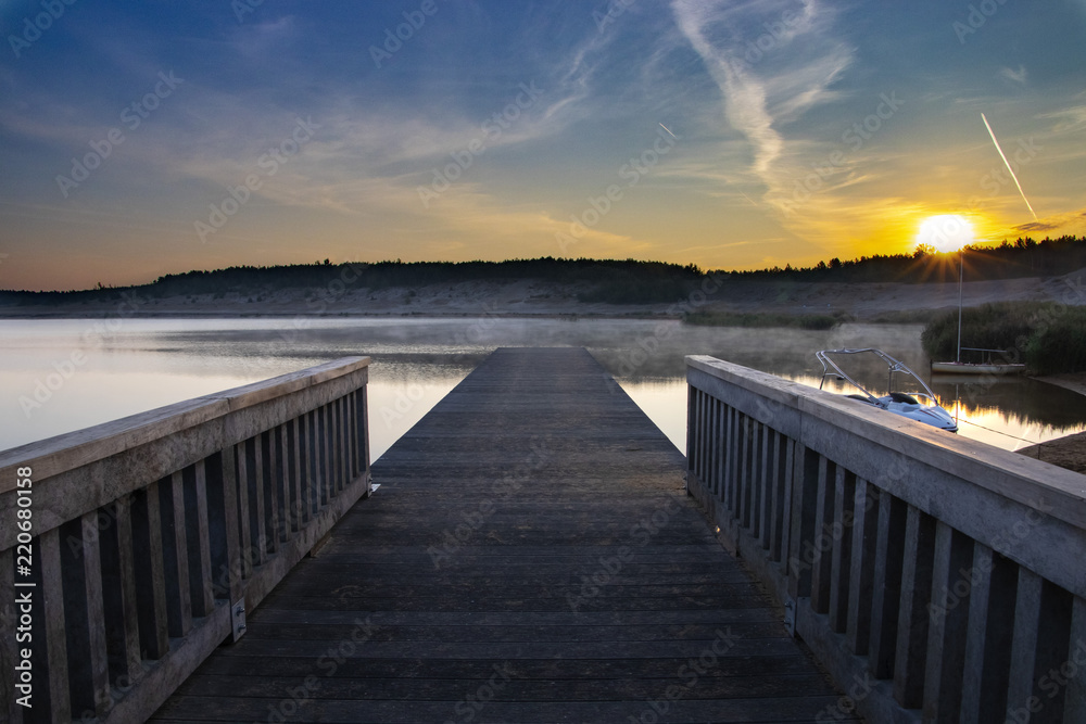 A wooden pier is standing on a lake. It is morning and sunrise shining orange, yellow and red lights. The pier is situated on a sandy beach. Morning mist is o the lake.