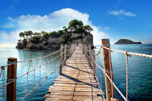 The wooden bridge overlooking the sea leads to an island with palm trees. It's a rope bridge. It is located in Zakynthos, Greece.