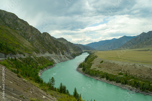 The turquoise river of the Katun in the mountains of the Altai among the rocks and green fields with derevy and grass in the autumn with a white sky and clouds