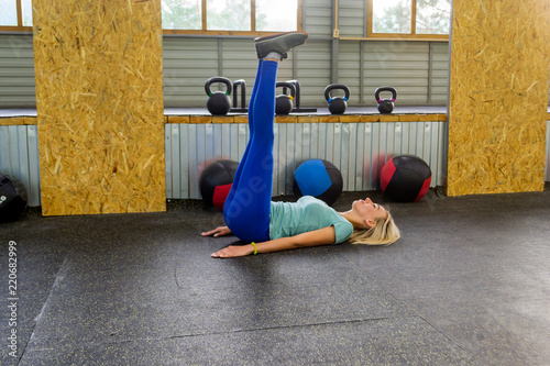 Blonde beatifull woman is making a warm-up lying on the floor raising up legs in the right angle, resting his palms and shoulder blades in the gym on the background of sports equipment photo