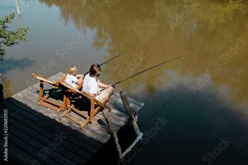Fotografie A young dark-haired man and a blond boy are sitting in recliners on the wooden pier with fishing rods and fishing