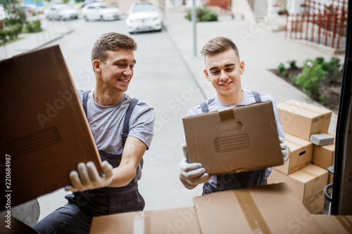 Two young handsome smiling movers wearing uniforms are unloading the van full of boxes. House move, mover service.