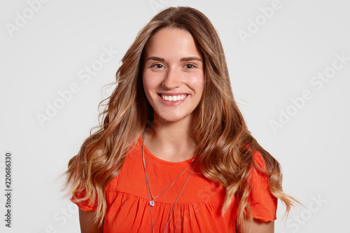 Close up portrait of smiling pleased European woman shows white teeth, has long hair, dressed in fashionable dress, being in good mood, isolated over studio wall. People and emotions concept © sementsova321