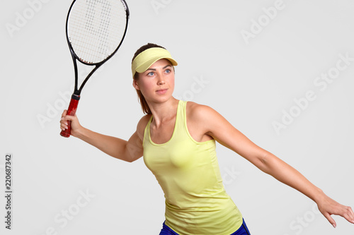Blue eyed pretty woman dressed in sportsclothes, ready to hit ball, has concentrated look into distance, likes playing tennis, isolated over white background, stands in ready posture. Sport concept © sementsova321