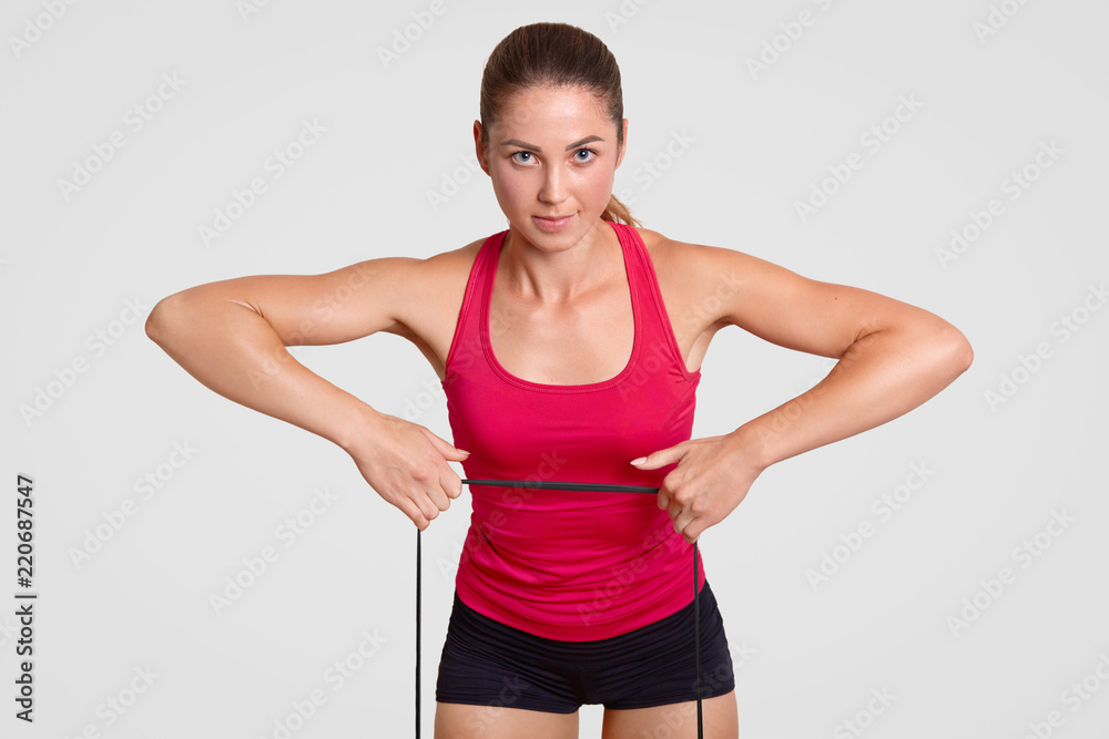 Strong healthy young fitness woman does stretching exercise with fitness gum, dressed in sportswear, looks directly at camera, makes athletic exercises, poses indoor against white background Strong healthy young fitness woman does stretching exercise with fitness gum, dressed in sportswear, looks directly at camera, makes athletic exercises, poses indoor against white background