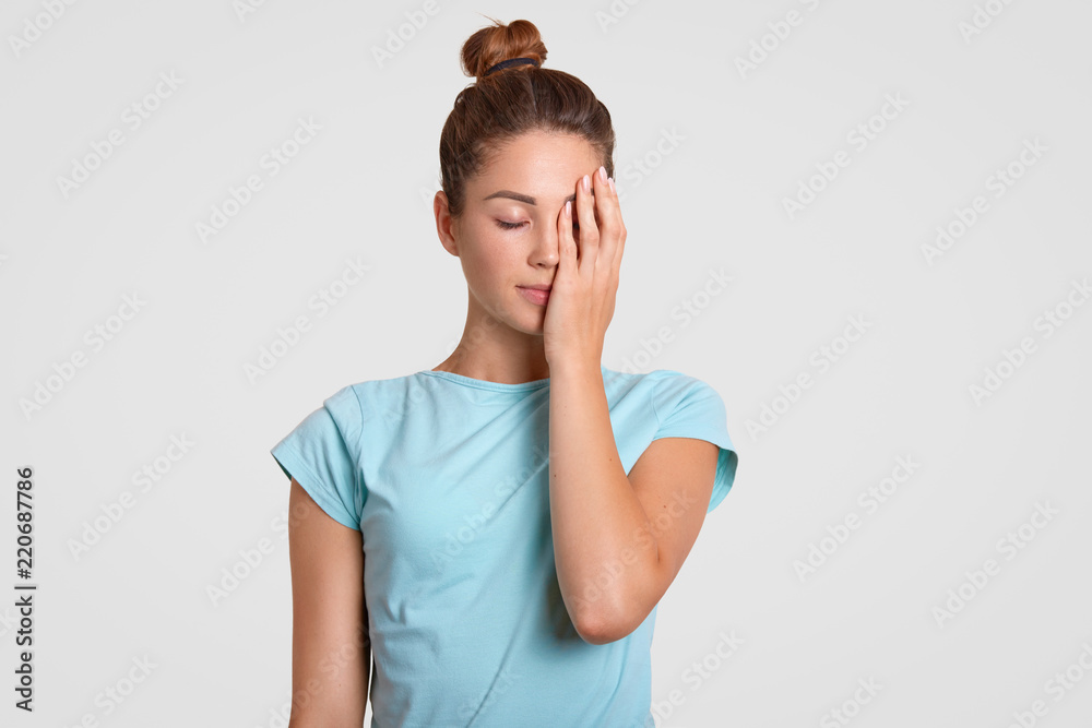 Indoor shot of tired sportswoman covers face with hand, dressed in casual t shirt, keeps eyes shut, tries to concentrated. Female fitness trainer feels exhausted after aerobic trainings, isolated Indoor shot of tired sportswoman covers face with hand, dressed in casual t shirt, keeps eyes shut, tries to concentrated. Female fitness trainer feels exhausted after aerobic trainings, isolated