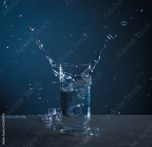 Ice splashes into glass of water on blue background
