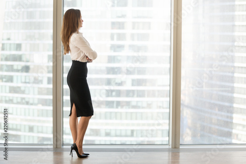 Successful businesswoman standing looking out of window enjoying city view and contemplating, confident rich woman leader thinking of business success and future vision planning new goals, copy space