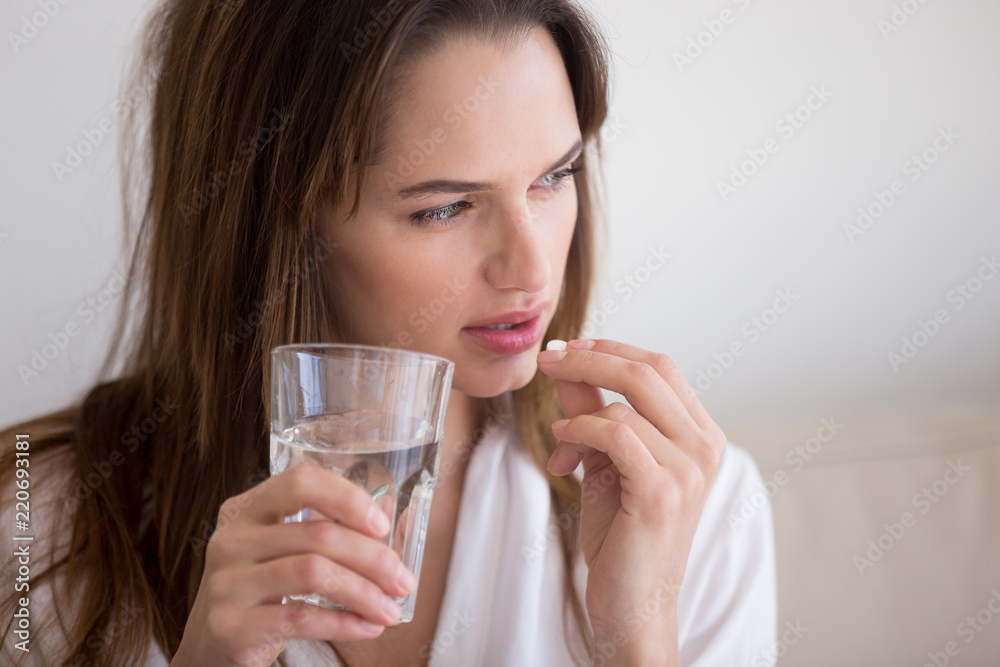 Doubtful sick ill young woman holding pill and glass of water taking painkiller medicine drugs to relieve headache pain, worried about side effects of antidepressant or emergency contraceptive meds - obrazy, fototapety, plakaty 