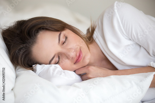 Young woman with beautiful face sleeping well on white cotton sheets and soft pillow lying asleep in comfortable cozy bed at home or hotel enjoying healthy nap resting enough for good relaxation.