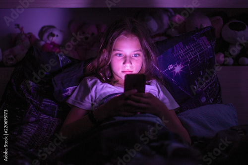Little girl lying under a blanket looking at the smartphone at night.