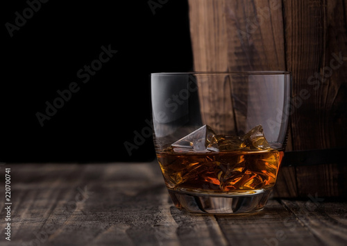 Glass of whiskey with ice cubes next to wooden barrel. Cognac brandy drink