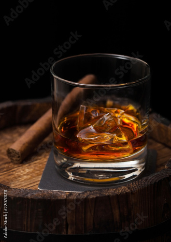 Glass of whiskey with ice cubes and cigar on top of wooden barrel. Cognac brandy drink