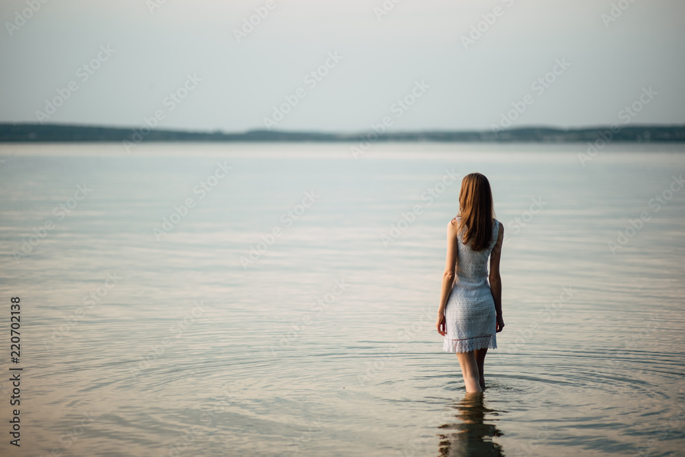Woman in summer dress standing on seashore and looking at horizon. Young beautiful girl standing in water