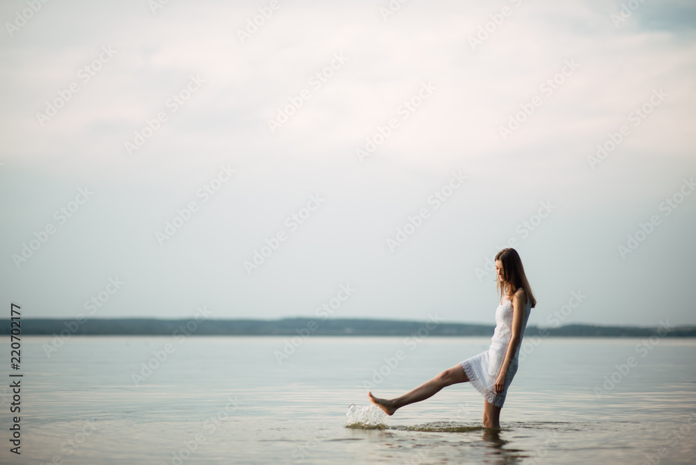 Woman in summer dress standing on seashore and looking at horizon. Young beautiful girl standing in water