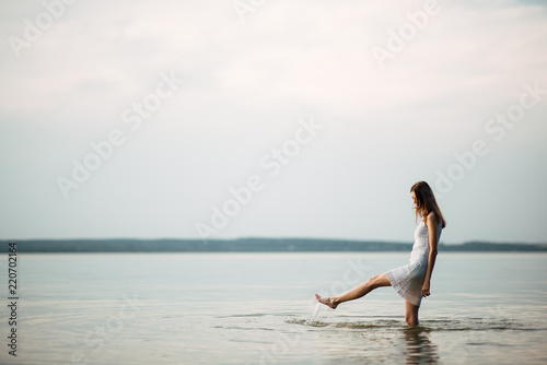 Woman in summer dress standing on seashore and looking at horizon. Young beautiful girl standing in water