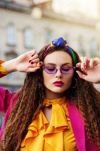 Outdoor close up fashion portrait of young beautiful woman wearing trendy violet sunglasses, colorful headband, circle hoop earrings, yellow blouse, pink blazer, posing in street of european city. 
