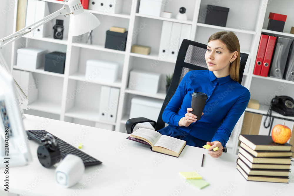 A young girl sits at a computer desk and holds a glass with coffee and a yellow marker in her hand. Before the girl lies an open book.
