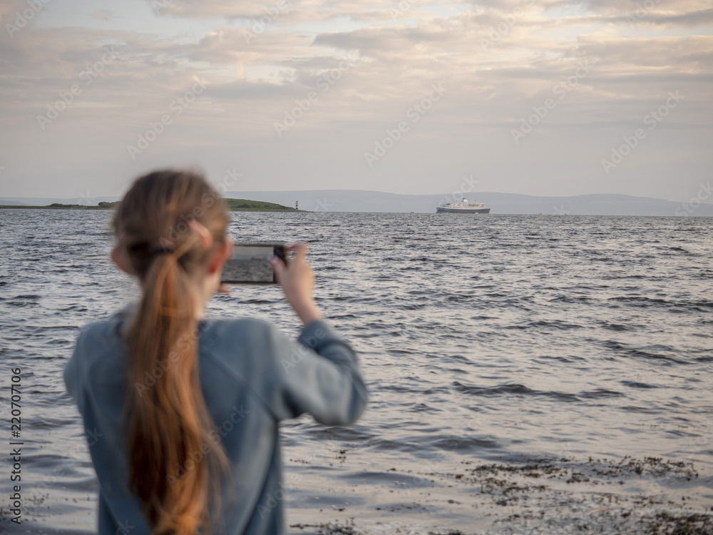 Girl using her smart phone camera to take a picture of  cruise liners to share it with her friends. The girl is out of focus,