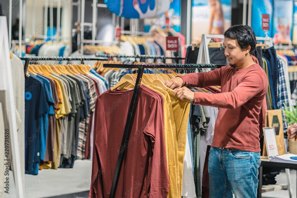 Obraz premium Smart asian man with beard choosing clothes in clothing store at shopping center, looking colorful t-sshirts , Fashion and Consumerism Concept.