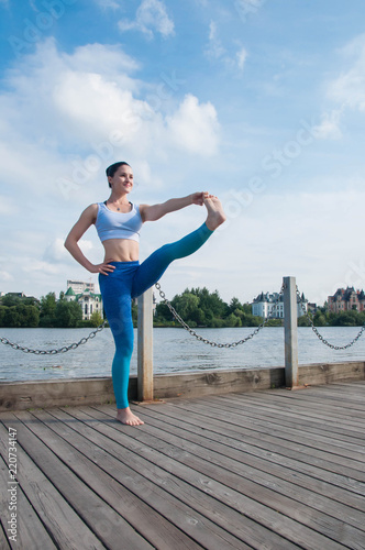 Young pretty fit and slim woman practices yoga and meditation on the embankment of the river