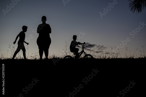 Silhouette of a boy walking, another boy on a bicycle and a woman - outdoors © Mateus