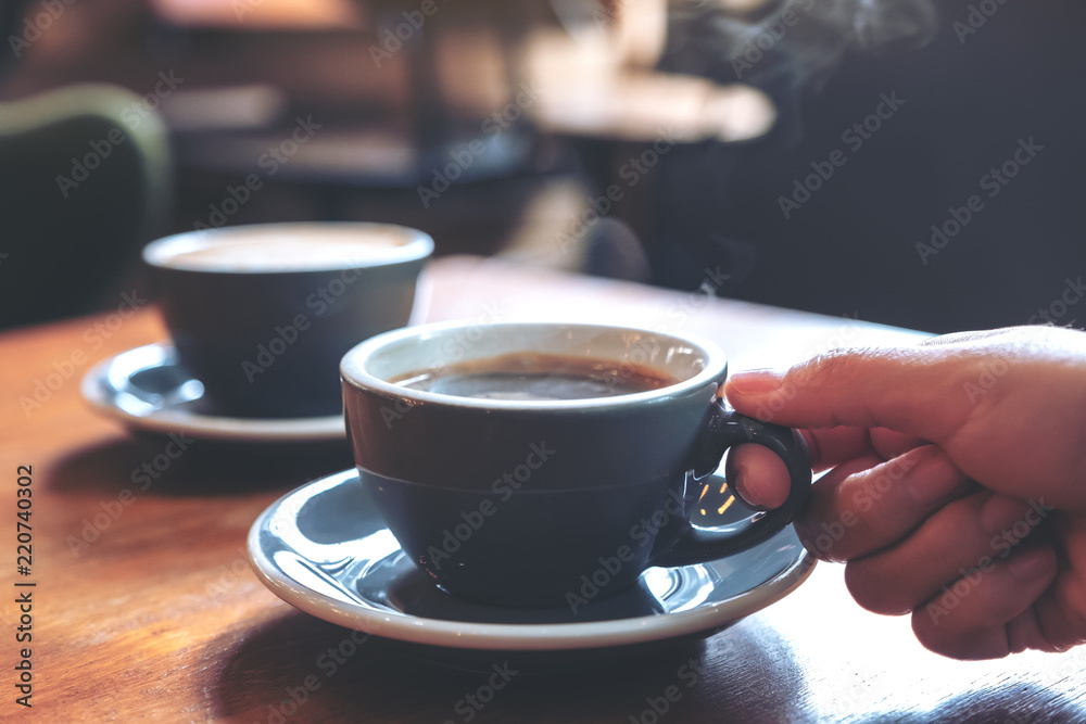 Fototapeta Closeup image of a hand holding a blue cup of hot coffee with smoke on wooden table in cafe